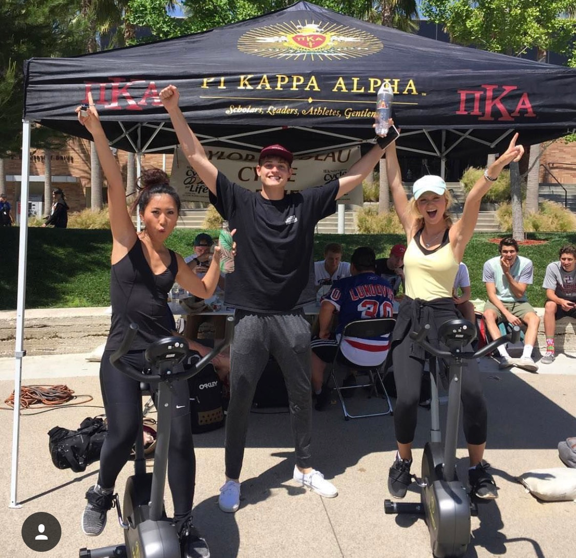 Two Young Women Cycle In Front of a Pi Kappa Alpha Popup Tent Photograph