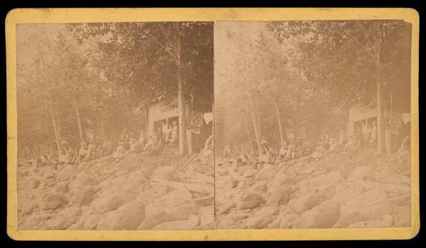 [Men, women, and children gathered in front of a house in the woods]