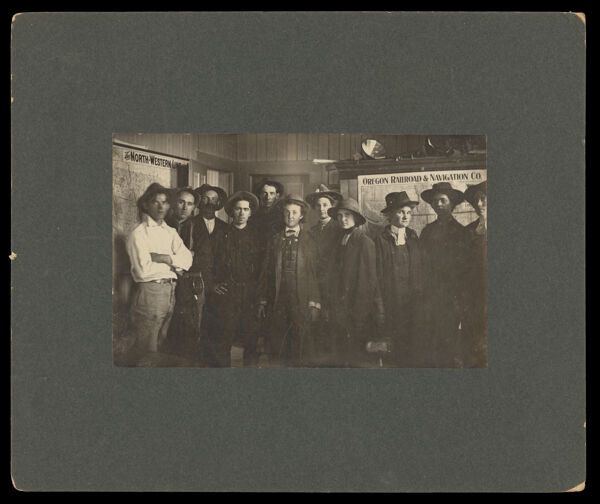 [photograph of 11 men and women standing in a railroad office, in front of a map for the North-Western Line and a map for the Oregon Railroad & Navigation Co.]