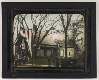 [People around house with American flag with portrait of man in center]