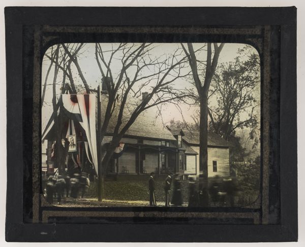 [People around house with American flag with portrait of man in center]