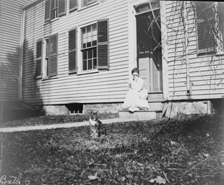Frances seated on steps in rear parsonage lot. Ted (cat) in grass near walk.