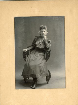 Woman in dark dress seated in a chair with hand under chin