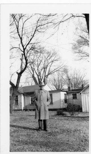 Man in light colored overcoat and hat standing near a light.colored house