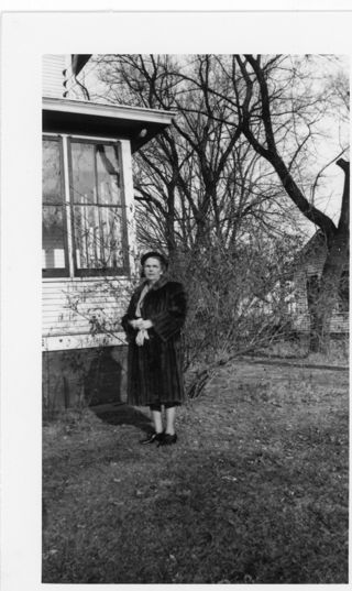 Woman in a dark colored fur coat standing next to a house
