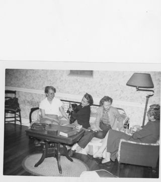 B&W photograph of 4 women seated in a living room, one holding a black cat and the other a dog in her lap.