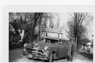 Man in suit, light colored overcoat and hat standing next to a 1940 or 1950 Packard car. No leaves on vegitation Man in suit, light colored overcoat and hat standing outside next to a 1940 or 1950 Packard car. No leaves on vegitation
